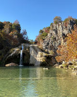A small waterfall flows into a clear pool surrounded by rocky cliffs and autumn foliage under a blue sky.