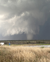 A person in a blue jacket stands in a grassy area observing a large tornado forming under a dark, turbulent sky.