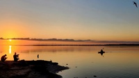Artist: Dominic Burch. 17 by 21-inch framed photograph. The photograph includes figures on a coastline of still water. Two of the figures are on the shore, another is paddling a craft in the water nearby. The sun is near the horizon and casts the land and figures in shadow. A thin line of clouds partially covers the sun and runs parallel to the horizon.