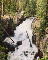 A waterfall emerges from a tree covered stone cliff. Vegetation grows down the rock on either side of the falls. The water and bright sunlight create rainbows at the base of the falls where the water splashes on the rocks below.