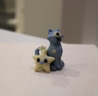 A blue cat-like sculpture sits next to a yellow star-shaped figure on a light surface.
