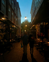 A couple walks hand-in-hand down a dimly lit alley flanked by buildings, with outdoor café seating and bicycles parked along the sides.