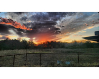 A sunset over a field, featuring dramatic clouds in shades of gray, orange, and blue, with a rusted metal fence in the foreground.