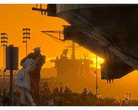 A statue of a sailor kissing a woman in a white dress is silhouetted against an orange sunset, with a ship and people in the background.