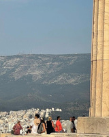 Tourists near ancient stone columns with a city and mountains in the background.