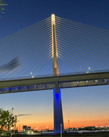 Suspension bridge at dusk with a lit support tower and blue lights, set against a gradient sky transitioning from blue to orange.