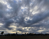 Dramatic low clouds hang heavy over houses and trees. Patches of blue sky and sunlight can be seen through the clouds.