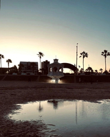 A beach at sunset featuring a lit gazebo, palm trees, and a shallow reflective pool in the foreground, with modern buildings in the background.