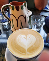A cappuccino with heart-shaped latte art, next to a patterned ceramic pitcher and a glass of water on a reflective table.