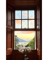 A window with an ornate wooden frame shows a colorful sunset over mountains and a lake, with white orchids in a pot in the foreground.