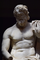 Image of a weathered bearded statue. The figure is bare chested with a drape at shoulder and waist. The nose and right arm of the statue are damaged.