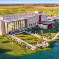 A vibrant image depicting a large, three-story building with a blue lake in front and many acres of green land behind it. 