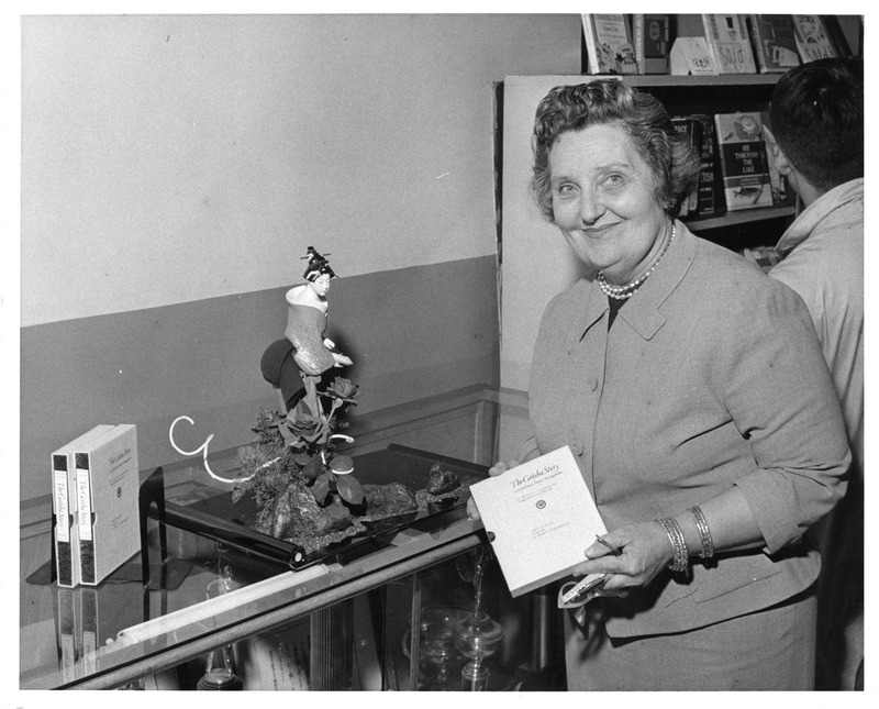 Woman standing next to a glass display case with a figurine and boxed items labeled "The Geisha."