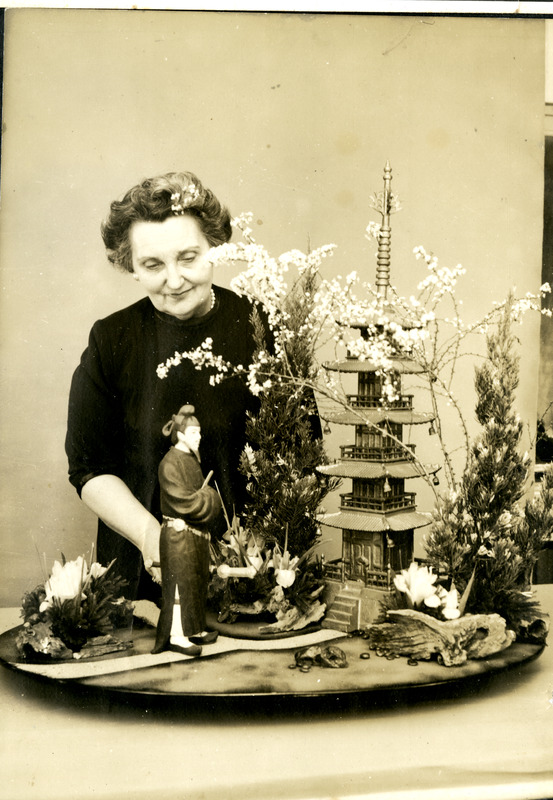 Older woman in a dark dress stands behind a detailed miniature Japanese scene with a figurine, trees, plants, and a pagoda.