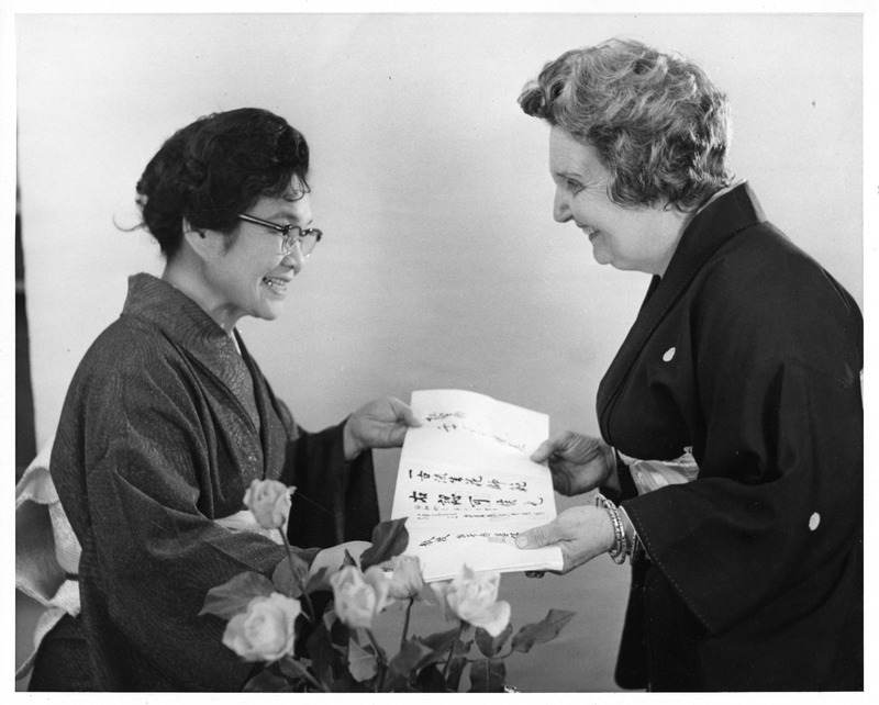 Two women in kimonos smiling at each other while holding a paper. A bouquet of roses is in the foreground.
