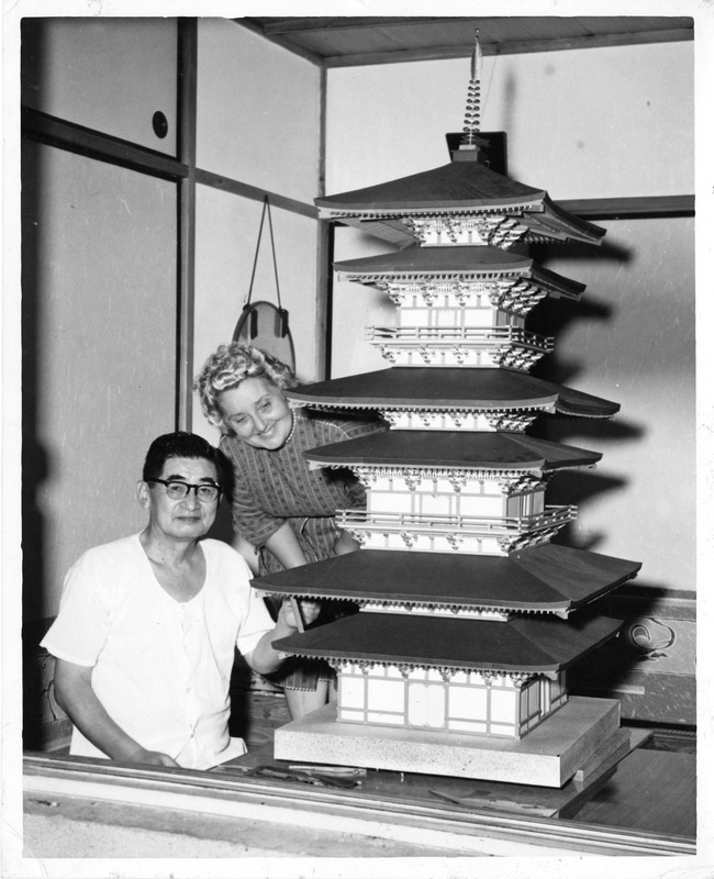 Two people beside a model of a multi-tiered pagoda indoors.