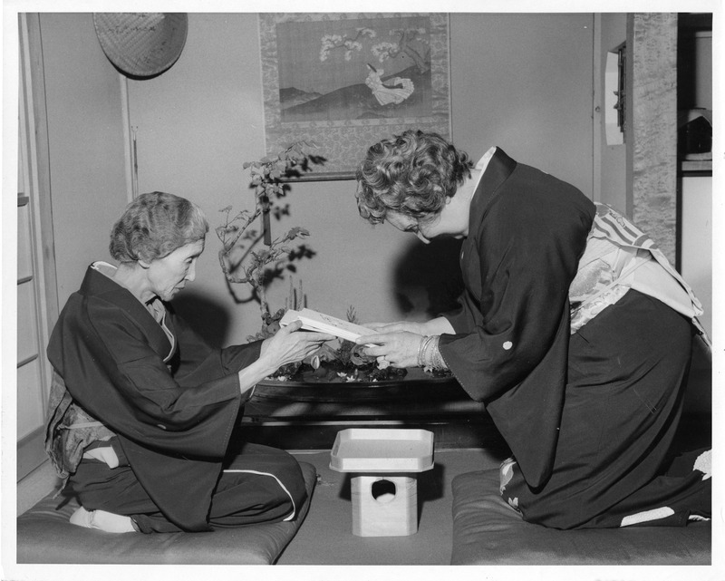 Two women in kimonos engage in a traditional Japanese tea ceremony, with a bonsai on a table between them.
