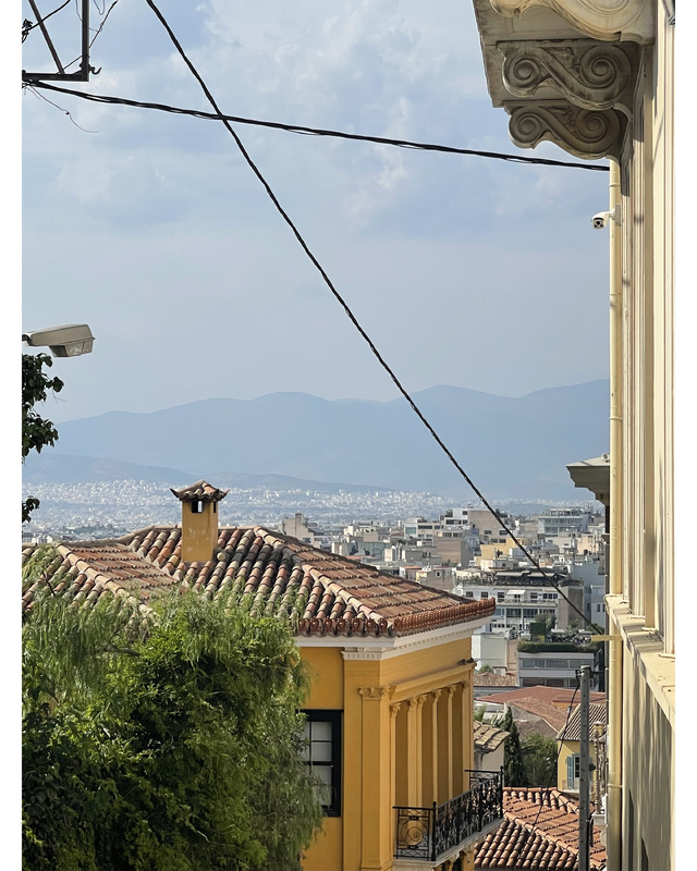 A view of rooftops and trees with a crowded city and mountains in the background. The sky is hazy and clouded with power lines running across the frame.