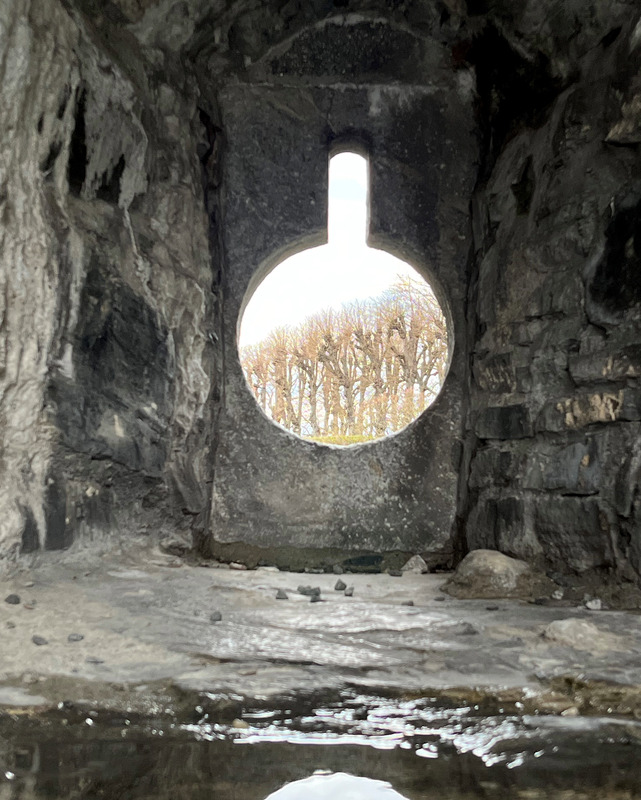 A line of bare winter trees  viewed from a keyhole shaped opening in a stone wall. Water is puddled in the foreground and reflects the light. coming through the hole.