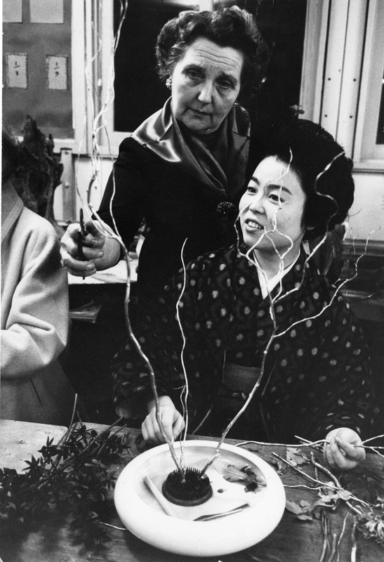 Two women arranging flowers, with branches and leaves on a table.