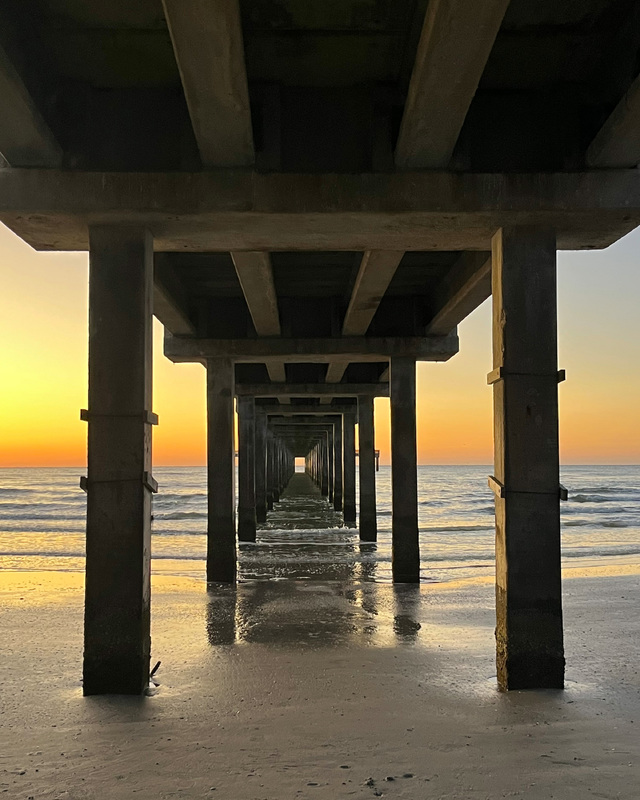Morning sunlight can be seen over incoming surf. The view is from the wet sand underneath a cement pier, looking out over the water.