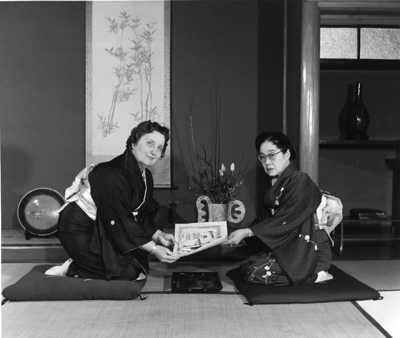 Two women in kimonos exchange a document while seated on cushions in a traditional Japanese room.