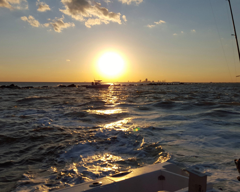 Sunset over a choppy sea with a silhouetted boat and distant city skyline.