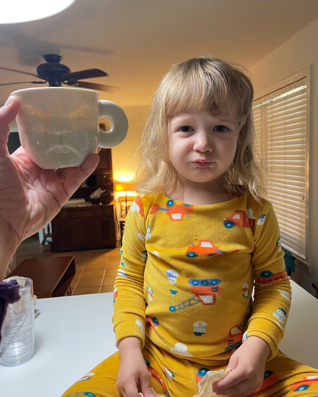 A child in yellow vehicle-themed pajamas sits on a countertop next to a hand holding a mug with a sculpted face.
