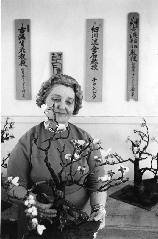 Woman holding a flower arrangement with Japanese calligraphy plaques on the wall behind her.