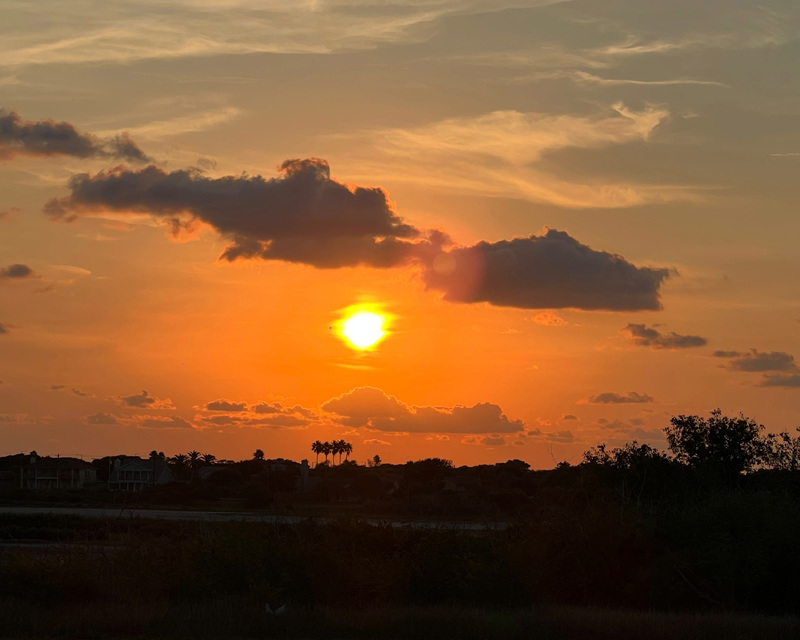 Sunset with an orange sky and scattered clouds above silhouetted trees and landscape.