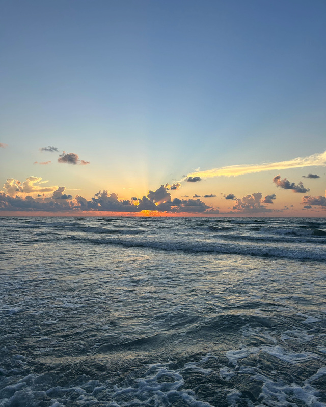 A sunrise over the ocean with gentle waves and scattered clouds.