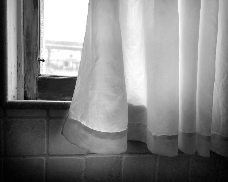 Black and white photo of a curtain next to a window with a blurred view outside and tiled wall beneath.