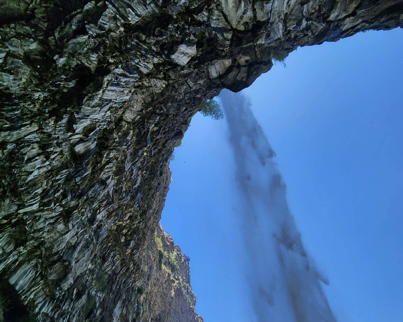 View of a waterfall from below, framed by textured rock formations against a blue sky.