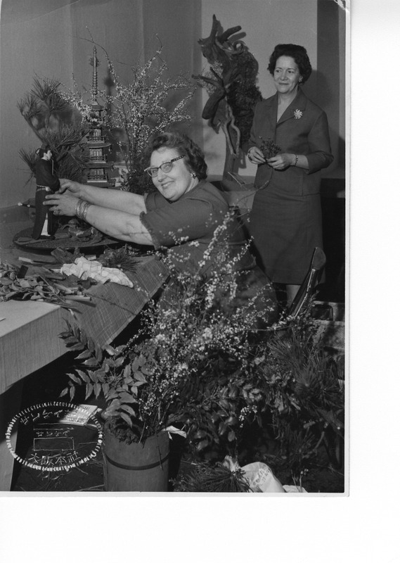 Two women arranging flowers and a small figurine on a table, surrounded by foliage and decorative elements.