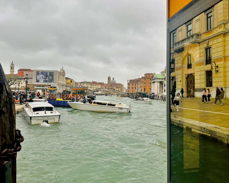 A busy canal scene with motorboats, historical buildings, and people by the water's edge. Reflection in glass shows pedestrians and an ornate building.