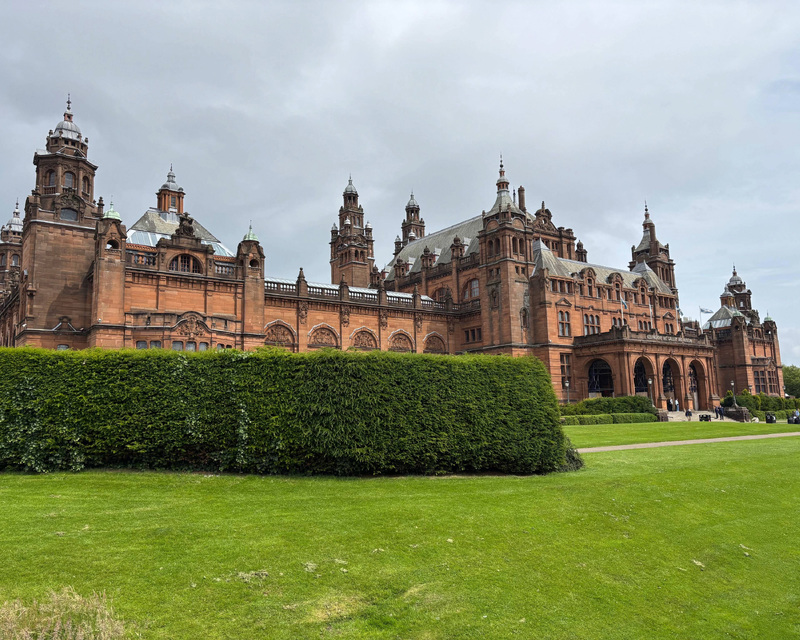 A historical building with reddish-brown stone, turrets, and ornate architecture, set behind a green lawn and hedge under an overcast sky.