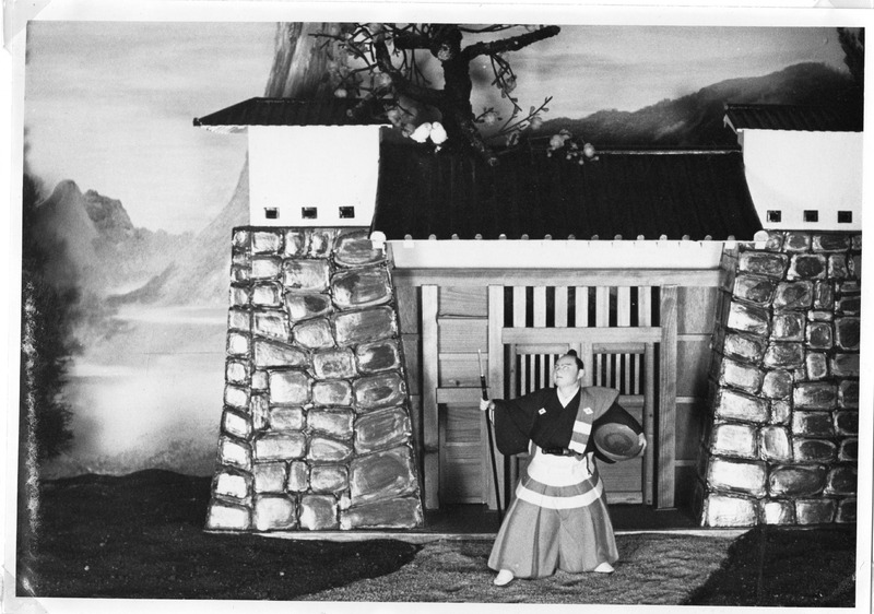 Performer in traditional costume stands by a Japanese-style gate with mountains in the background.