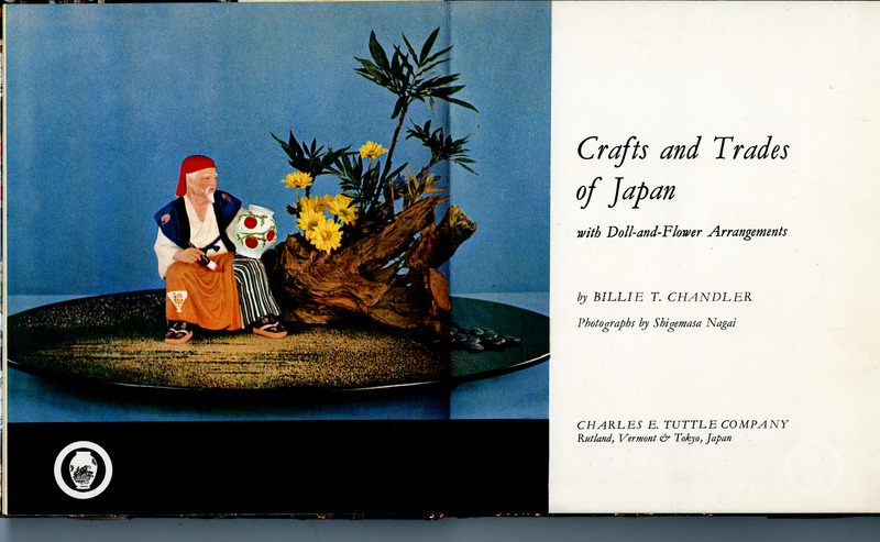 Cover of 'Crafts and Trades of Japan' featuring a doll dressed as a traditional Japanese tradesman seated beside a floral arrangement with yellow flowers and a wooden structure.