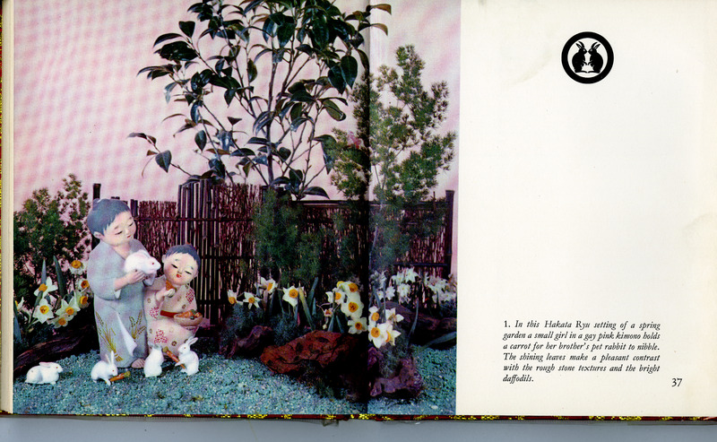 Spring garden scene with figurines of two children with rabbits, surrounded by daffodils and greenery.
Transcribed Text:
In this Hakata Ryu setting of a spring garden a small girl in a gay pink kimono holds a carrot for her brother's pet rabbit to nibble. The shining leaves make a pleasant contrast with the rough stone textures and the bright daffodils.