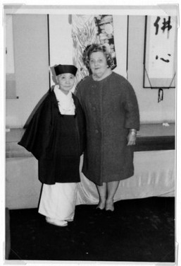 Two individuals in traditional and modern clothing stand in front of art-adorned walls.