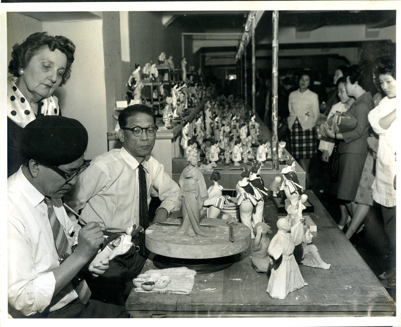 People working with and observing small sculptures in a workshop setting; sculptures and figures cover the tables.