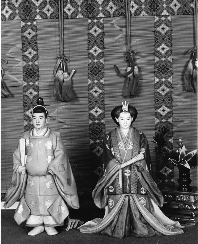 Two traditional Japanese dolls in front of a patterned backdrop with tassels.