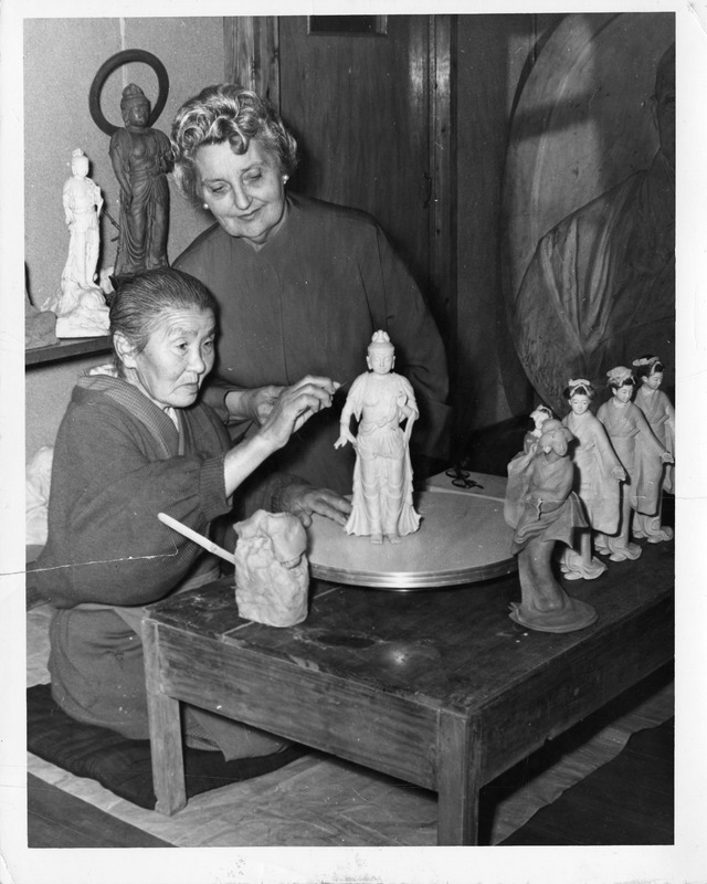 Two women sculpting figurines at a table with various clay statues.