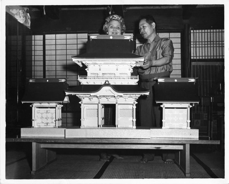 Two people look at a model of a traditional Japanese building with detailed roofs and panels on a table. Shoji screens are in the background.