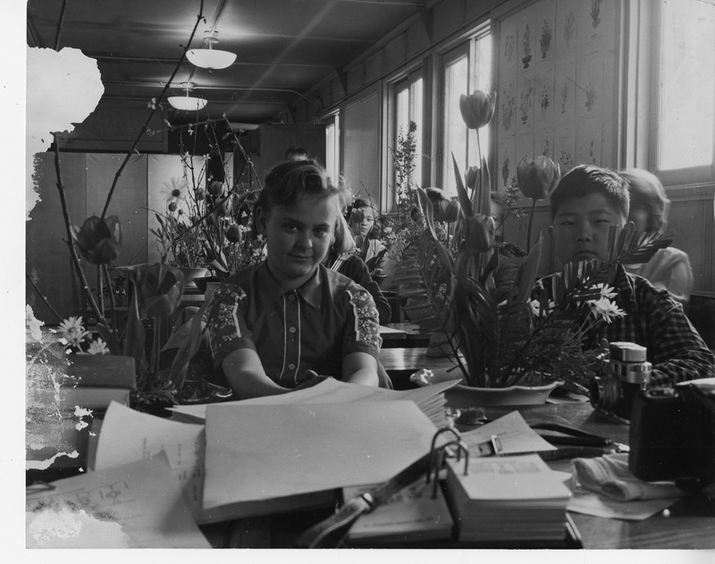 Students seated in a classroom with floral arrangements on desks; papers and camera in the foreground.