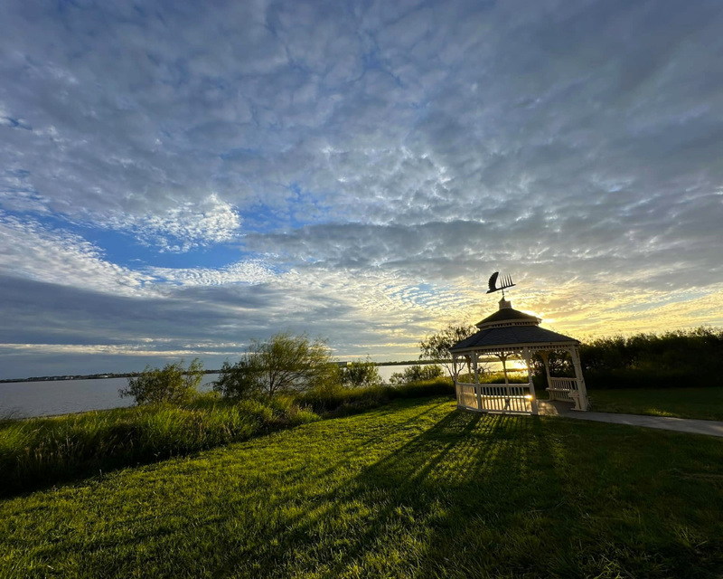 Gazebo on a grassy lawn near a lake at sunset with a cloudy sky.