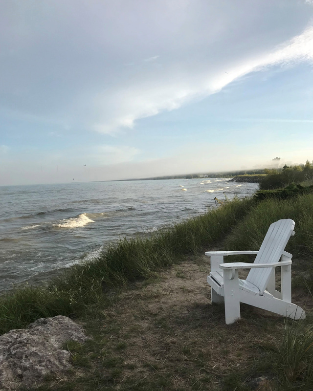 White Adirondack chair on a grassy lakeside with cloudy sky and gentle waves.