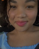 A close up portrait of a young person in a scooped neck light blue blouse. The person is smiling at the photographer, wearing pink lip gloss and large silver hoop earrings. One curling tendril of dark brown hair falls at the corner of their right eye, dropping down to chin level.