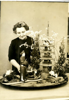Older woman in a dark dress stands behind a detailed miniature Japanese scene with a figurine, trees, plants, and a pagoda.