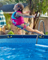 A young swimmer jumps into a raised swimming pool. The swimmer is wearing a life jacket and googles. The swimmer is caught mid-jump with back arched, arms wide and behind, with legs pointed behind. Parts of a building, umbrella, plants, trees, and fence are visible in the background.